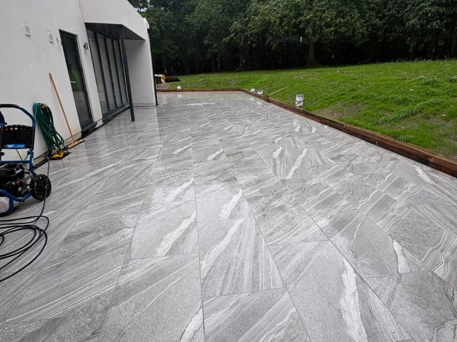 Grey veined porcelain patio laid alongside a white rendered house, bordered by lawn and trees