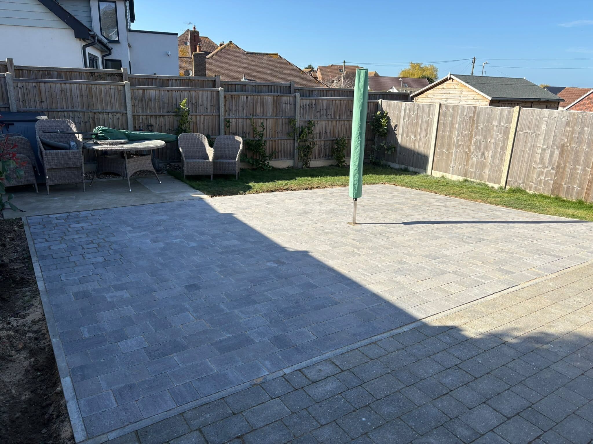 New grey block paved rear patio with rattan garden furniture and a parasol, framed by close-board fencing