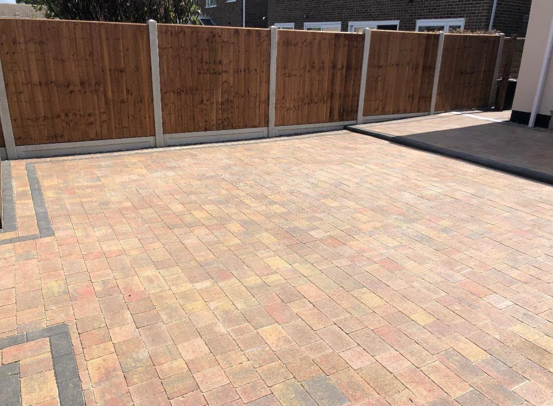 Buff and autumn-toned herringbone block paving with a charcoal soldier-course border beside a fence