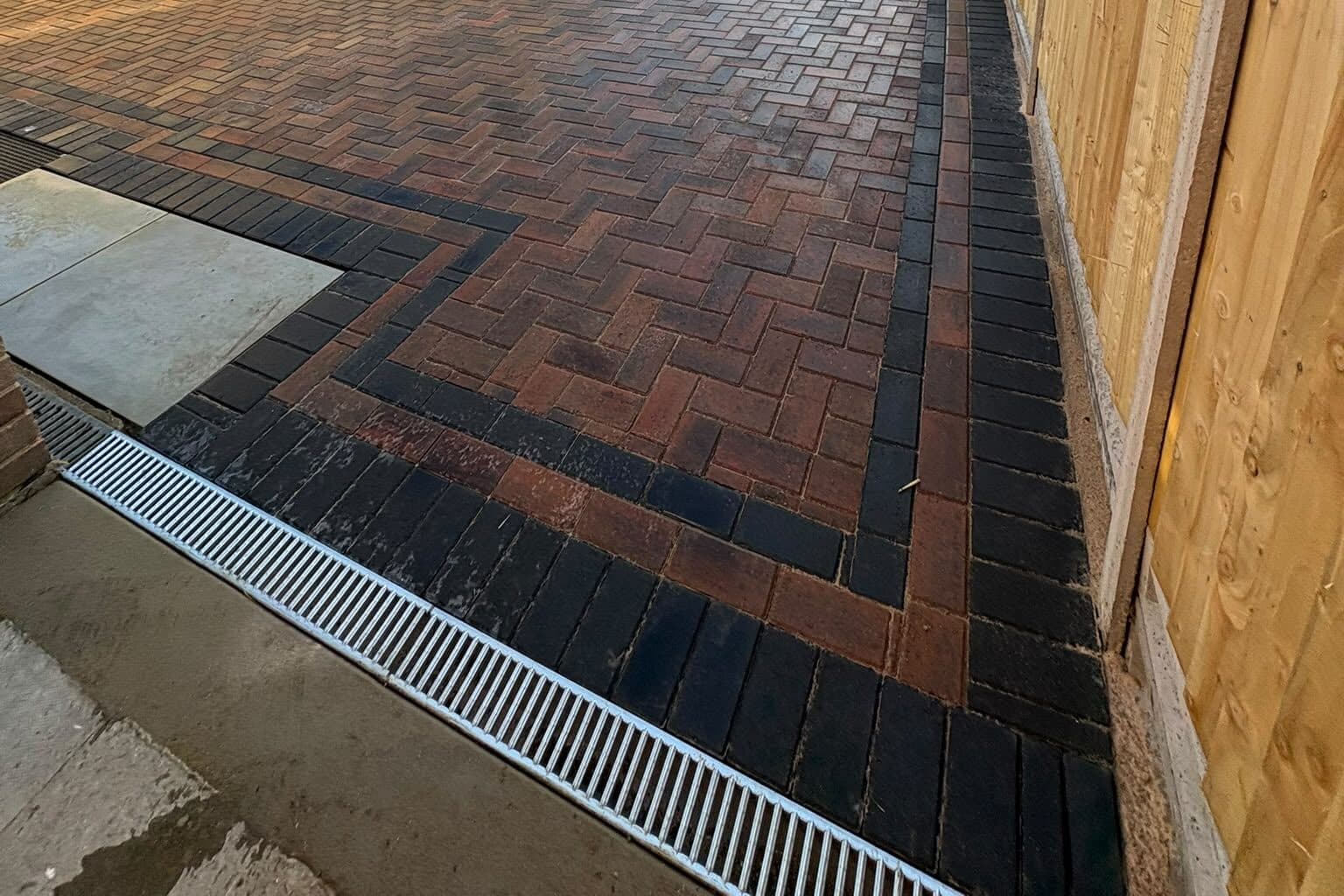 Close view of red and black herringbone block paving framed by a black border and drainage channel